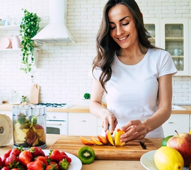 Person in well-lit, white kitchen, slicing a peach and surrounded by fresh fruit.