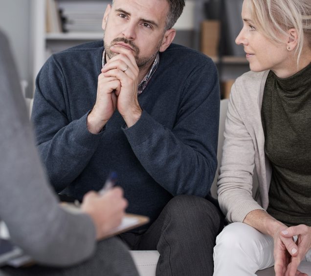 Couple sitting at a therapy session with a man in a deep gaze, while his female partner looks at him with hands folded.