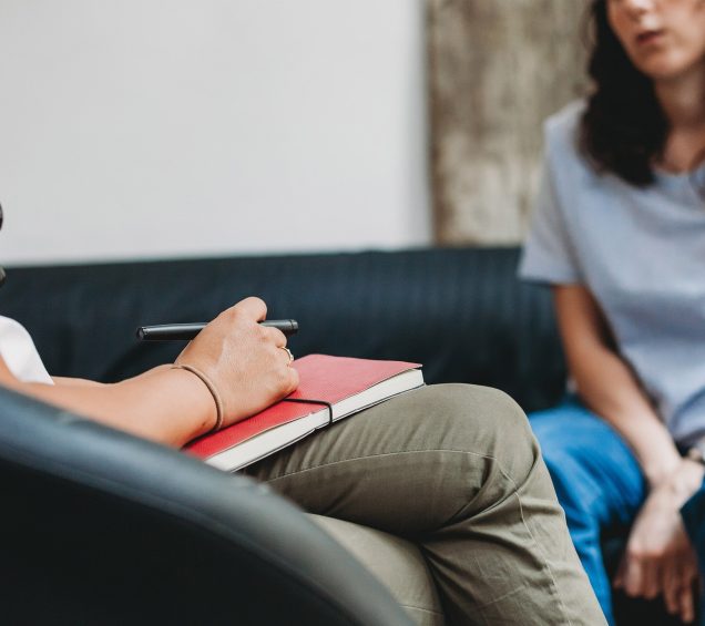 Female doctor with a red notebook and pen talking to a female patient in consulting session.