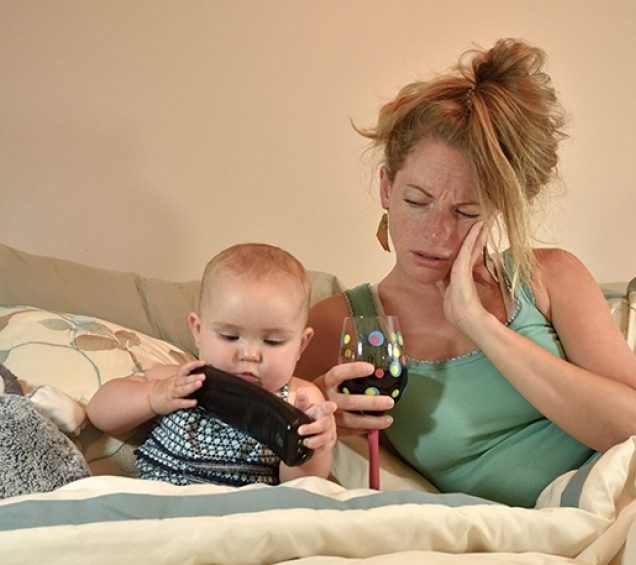 Distraught looking mother laying in bed with her baby, holding a glass of wine.