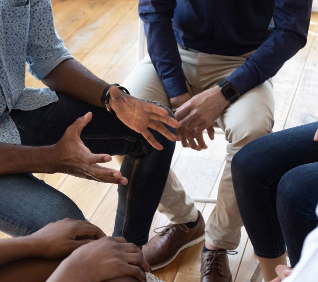 Group of people sitting in a circle with the focus on someone expressively talking with their hands open.