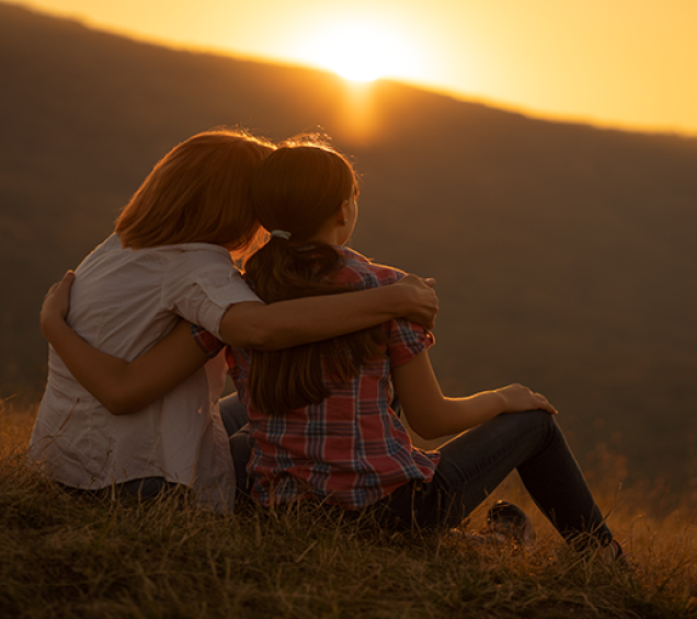 Parent and adult child sitting in a field at sunset with their arms around each other.