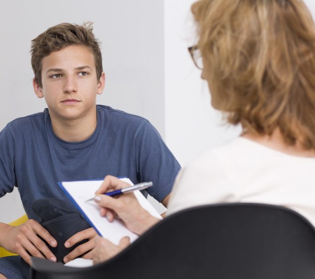 Young male sitting in a room with female counselor that is taking notes on a clipboard with pen.
