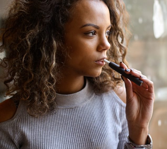 Young teen vaping with e-cigarette while looking out a window.