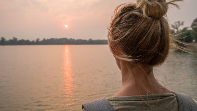 Woman with back turned overlooking a sunset over a lake.