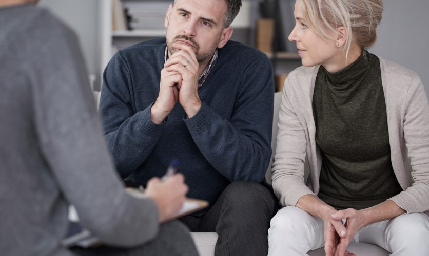 Couple sitting at a therapy session with a man in a deep gaze, while his female partner looks at him with hands folded.