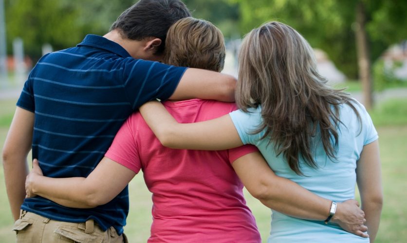 Family Comforting one another with their backs turned to the audience while outside at a park.