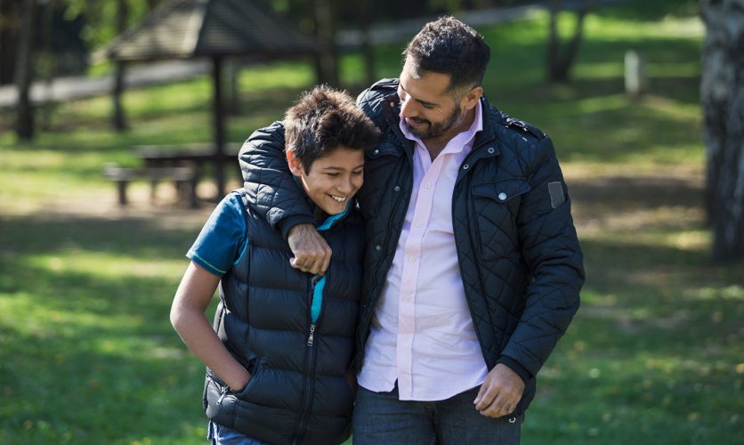 Father with arm around his son while walking in a green grass park.