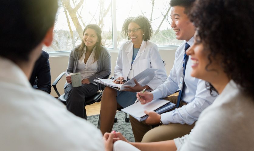 Group of physicians sitting indoors while holding notebooks and smiling.