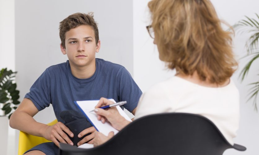 Young male sitting in a room with female counselor that is taking notes on a clipboard with pen.