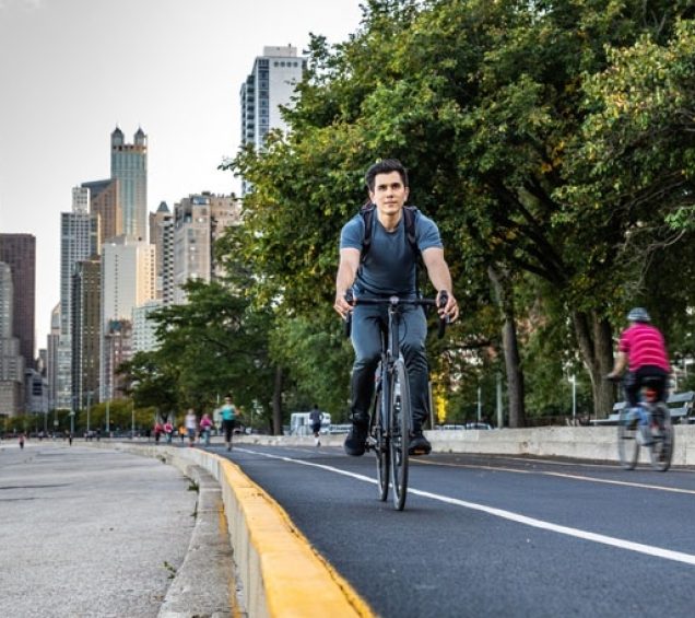 Young man riding a bike on a path with city skyline behind him.