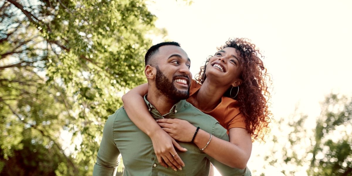 A smiling couple enjoys a playful moment outdoors, with the woman on the man’s back while they both look happy under the trees.
