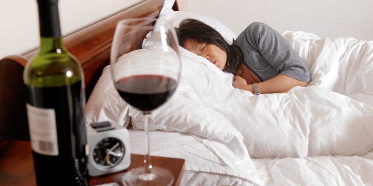 Woman lying in bed with a bottle, glass of wine, and pills on the nightstand.