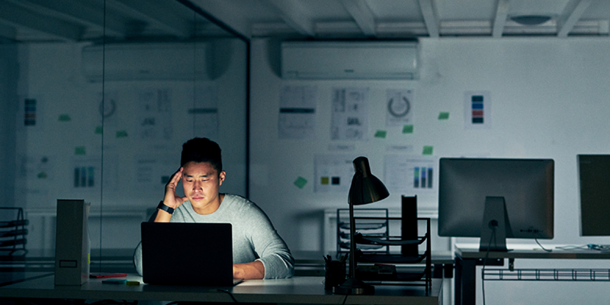 Man in a darkened and empty office, looking concerned and staring at a laptop screen.