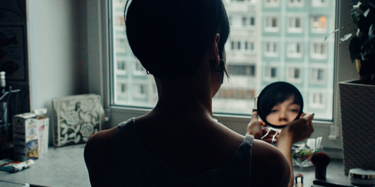Person at a window, holding a small mirror to look at their reflection.