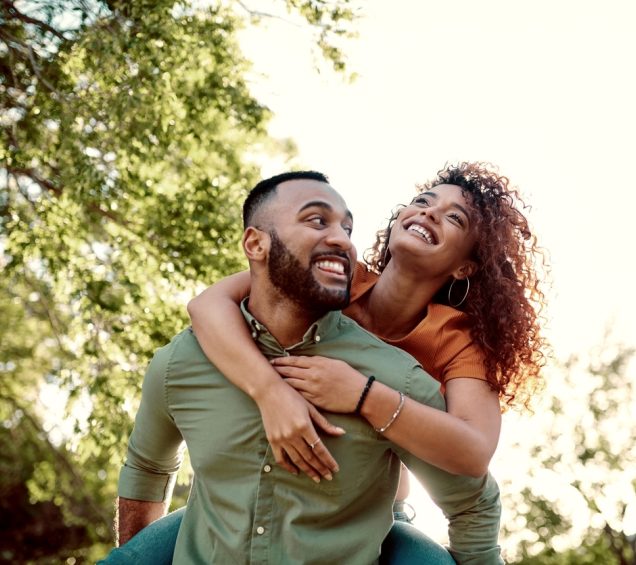 A smiling couple enjoys a playful moment outdoors, with the woman on the man’s back while they both look happy under the trees.