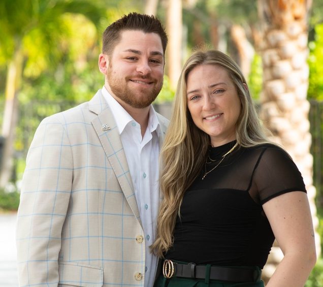 Couple standing close together if front of palm trees.