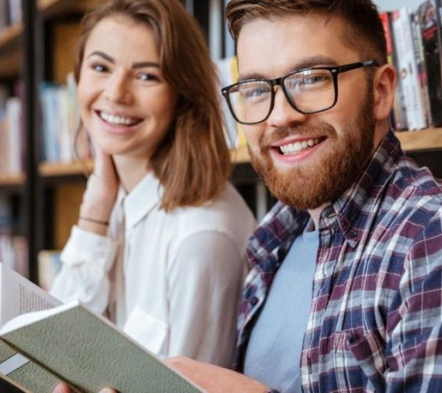 Two College students smiling inside a library while holding a book.