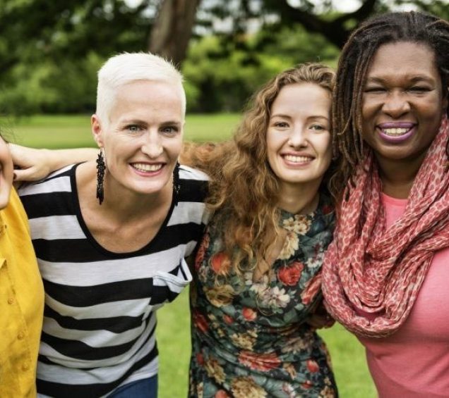 Diverse group of women smiling outdoors with their arms on each others shoulders.