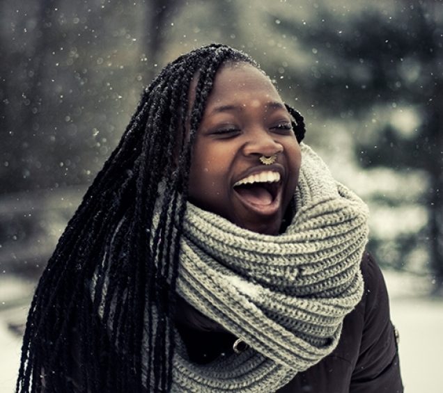 A woman outside in a gray scarf is smiling as snow is falling.