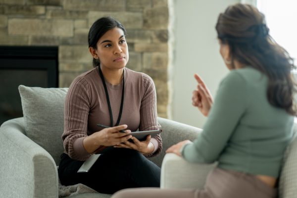 A therapist listens attentively and takes notes while a woman speaks during a counseling session in a comfortable office setting.